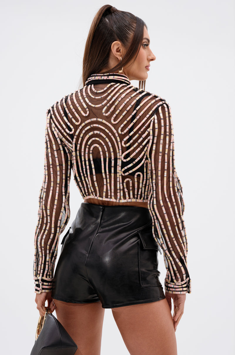 A woman with long brown hair in a high ponytail is wearing the EVA LONG SLEEVE BUTTON DOWN STRIPED SHIRT and black high-waisted leather shorts, holding a small handbag. She faces away, looking over her shoulder.