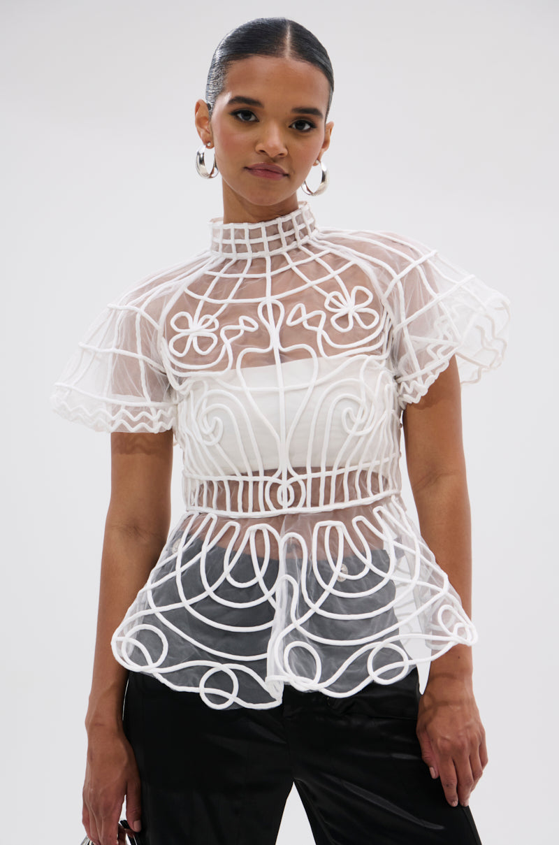 A woman stands against a plain light background wearing the DOLL FACE SHORT SLEEVE STRUCTURED BLOUSE—featuring sheer mesh, a high neck, ruffled sleeves, and intricate white embroidery over a white bandeau—paired with black pants.