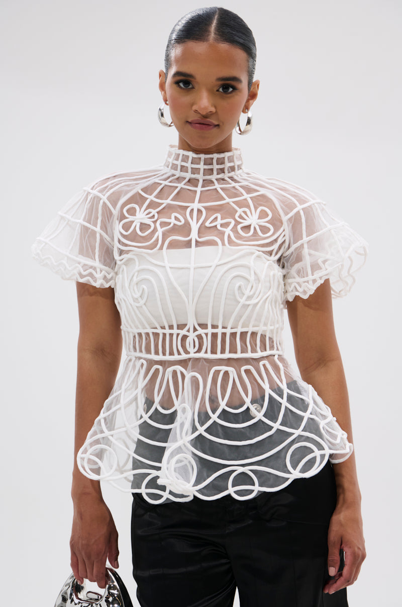 A woman wears the DOLL FACE SHORT SLEEVE STRUCTURED BLOUSE with black pants, accessorized with a silver handbag and large hoop earrings. Her hair is sleek and pulled back.