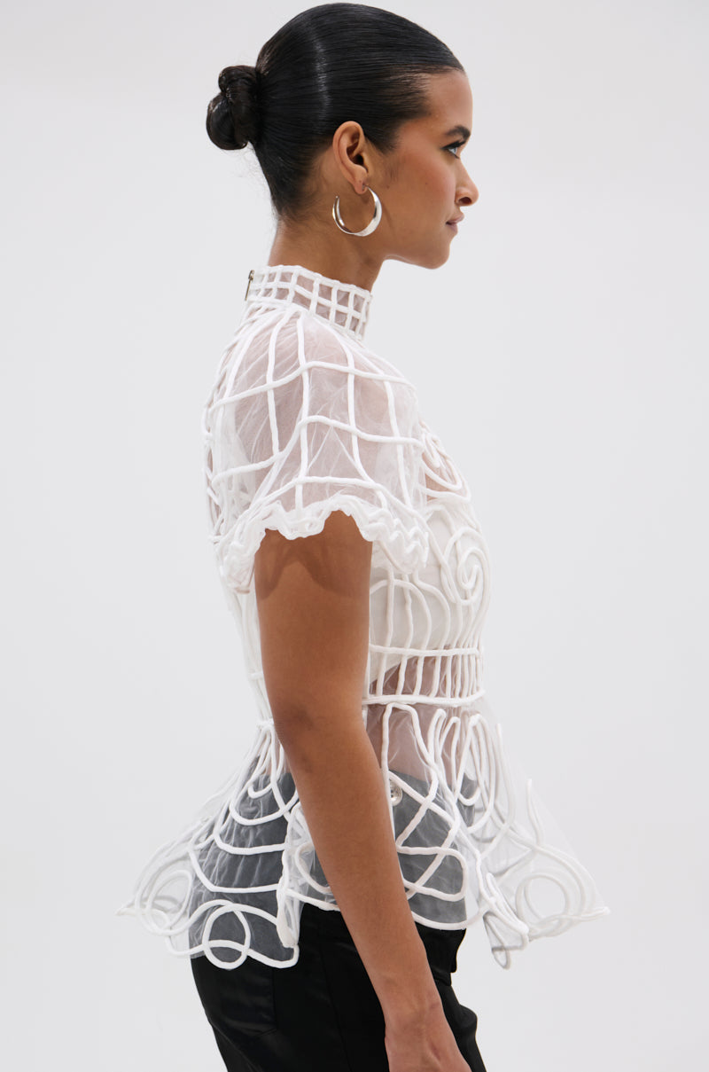 A woman with dark hair in a bun and large hoop earrings wears the DOLL FACE SHORT SLEEVE STRUCTURED BLOUSE with geometric patterns and a peplum hem, paired with black pants, standing in profile against a plain background.