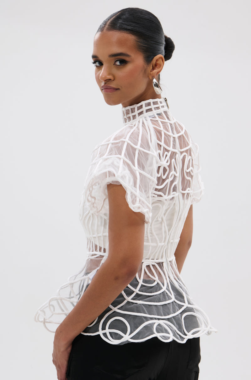 A woman with slicked-back hair poses in the DOLL FACE SHORT SLEEVE STRUCTURED BLOUSE, a sheer mesh top with geometric patterns, looking over her shoulder against a plain background.