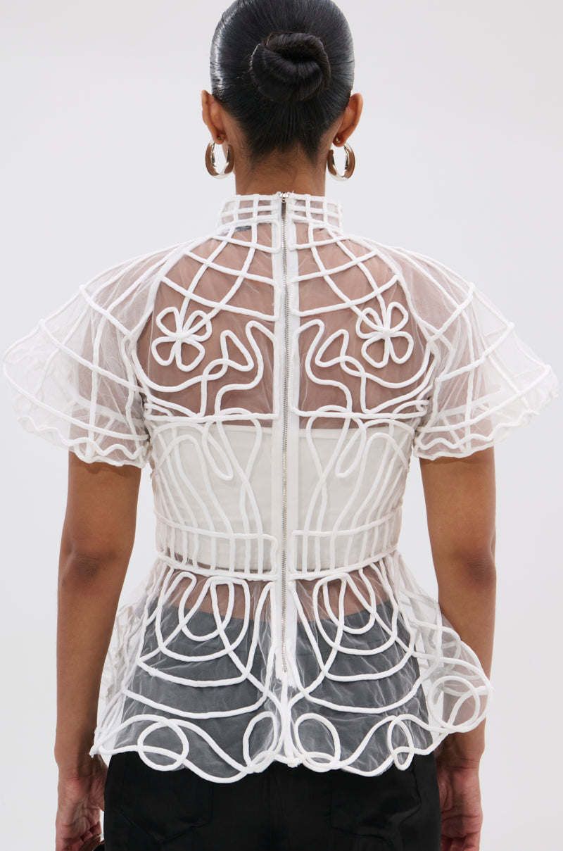 Seen from behind, a person wears the DOLL FACE SHORT SLEEVE STRUCTURED BLOUSE featuring sheer mesh fabric, geometric and floral embroidery, and a high collar. Their hair is in a bun and they're accessorized with large hoop earrings.