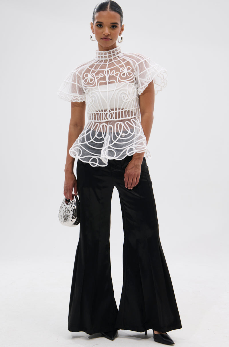 A woman stands confidently in the DOLL FACE SHORT SLEEVE STRUCTURED BLOUSE, featuring sheer mesh with swirling patterns and ruffled sleeves, styled with high-waisted black flared pants, black heels, and a small silver handbag.