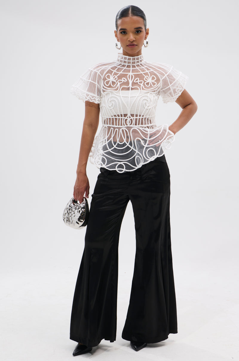 A woman stands confidently against a plain white background, wearing the DOLL FACE SHORT SLEEVE STRUCTURED BLOUSE with black wide-leg pants, black pointed shoes, and holding a small black-and-white patterned purse.