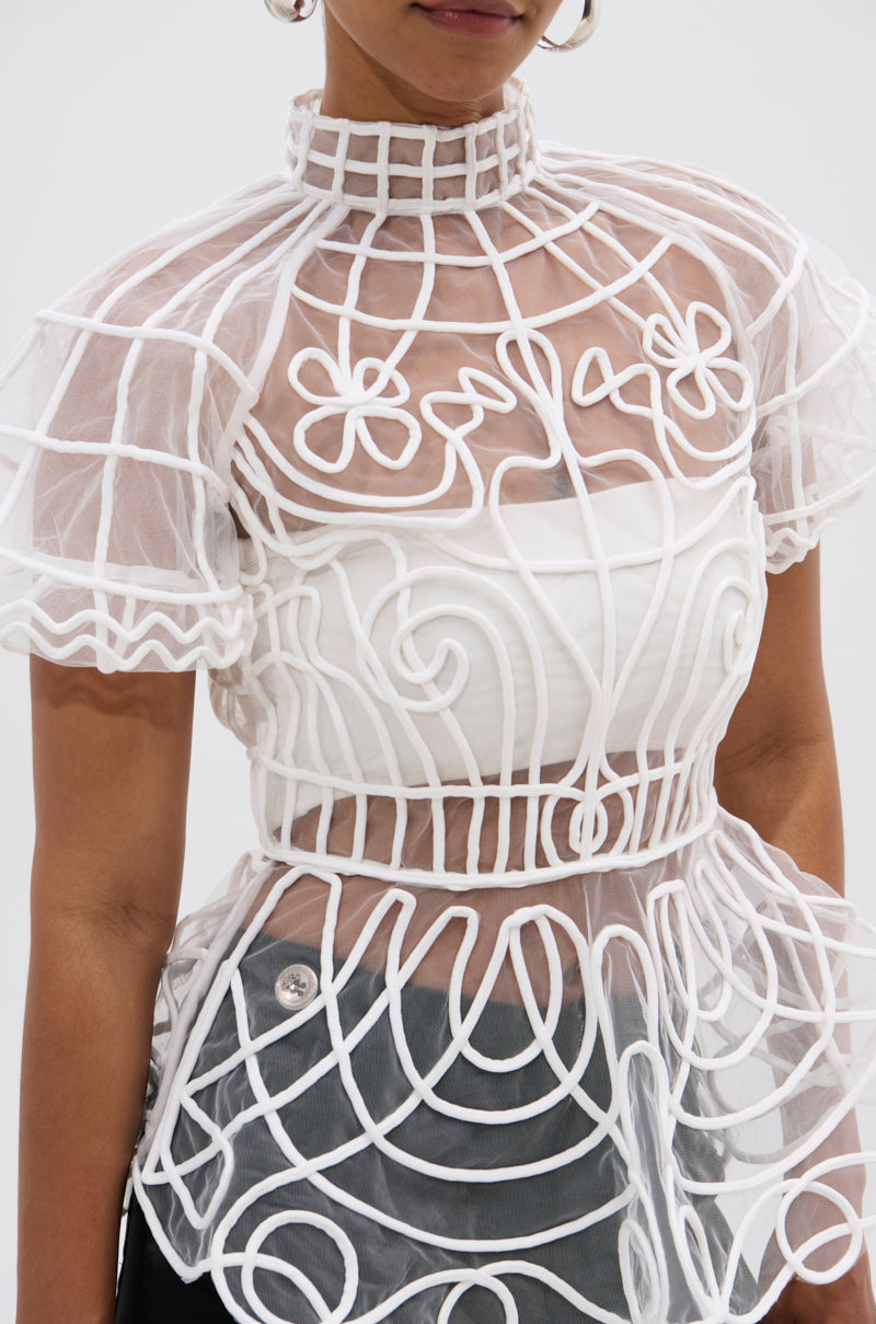 A person wears the DOLL FACE SHORT SLEEVE STRUCTURED BLOUSE, featuring sheer mesh with swirling floral embroidery, a high grid-patterned collar, and puffy sleeves over a white bandeau, paired with dark pants for an elegant look.