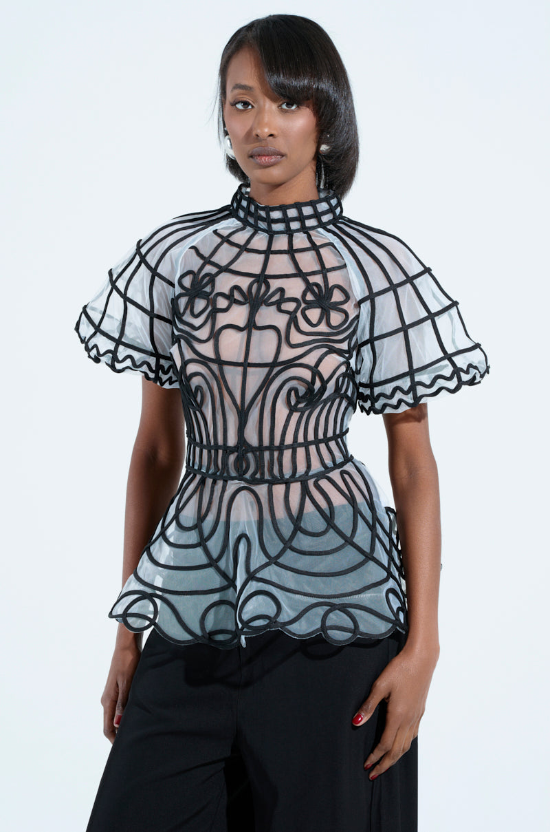A woman poses confidently against a plain background in the DOLL FACE SHORT SLEEVE STRUCTURED BLOUSE, which features intricate black patterns. Her straight hair complements her look, paired with black pants, conveying a poised demeanor.