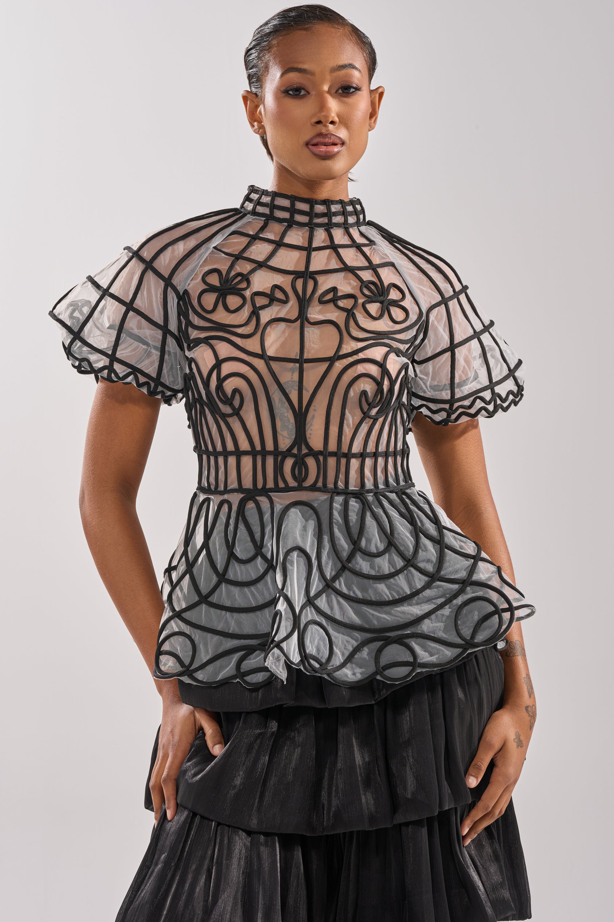 A woman with slicked-back hair and a confident expression models the DOLL FACE SHORT SLEEVE STRUCTURED BLOUSE, featuring sheer mesh, bold black piping, ruffled sleeves, a high collar, and a peplum silhouette, paired with a layered black skirt.