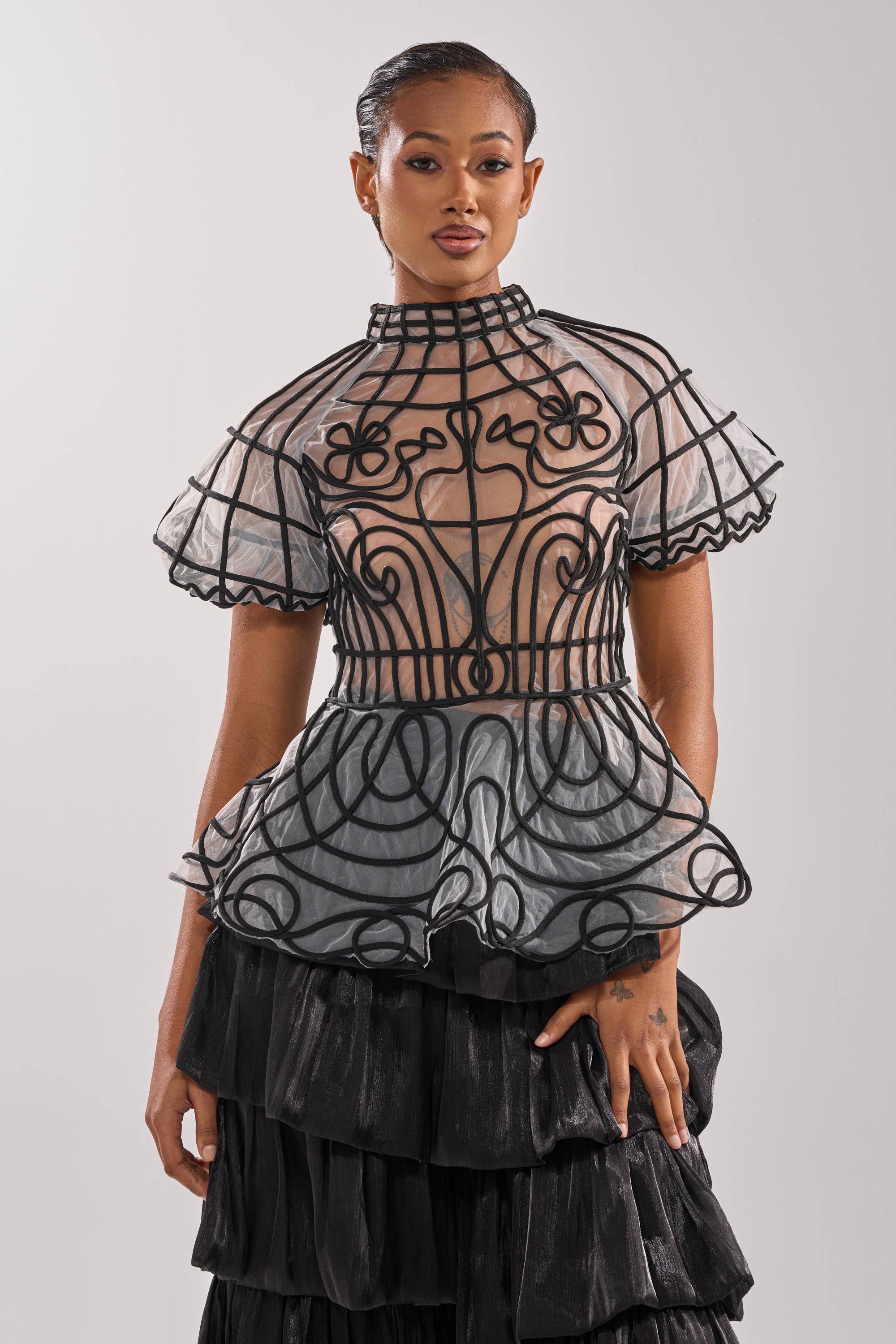 A woman models the DOLL FACE SHORT SLEEVE STRUCTURED BLOUSE, featuring sheer mesh with bold black geometric and floral designs, paired with a tiered black skirt. She stands against a light background with slicked-back hair.