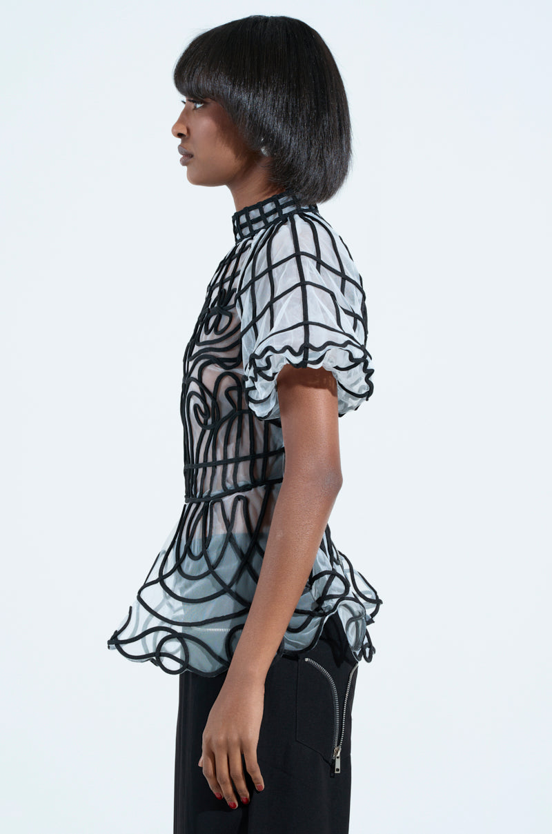 A person with medium-length hair stands in profile, wearing the DOLL FACE SHORT SLEEVE STRUCTURED BLOUSE. It features a sheer black geometric pattern, high collar, puffed sleeves, and flared peplum silhouette. A plain white background highlights the chic design.