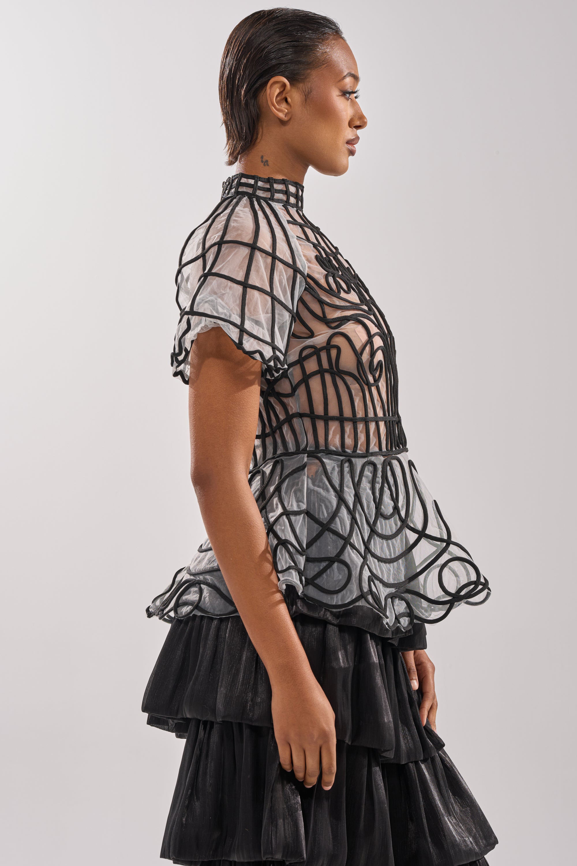 A woman stands in profile wearing the DOLL FACE SHORT SLEEVE STRUCTURED BLOUSE with a ruffled black skirt. Her hair is slicked back, and the background is plain light gray.