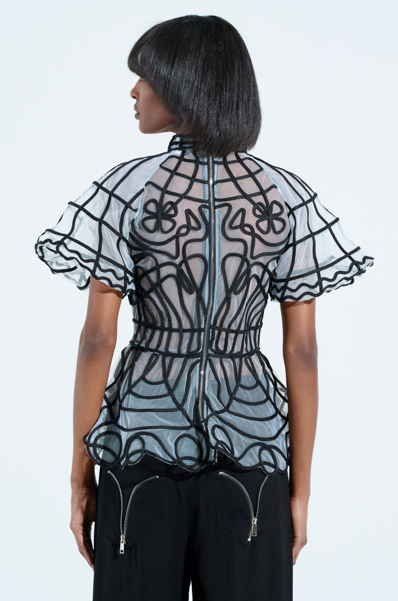 A person with short, straight black hair wears the DOLL FACE SHORT SLEEVE STRUCTURED BLOUSE with intricate sheer designs and ruffled sleeves, paired with black pants featuring zippers. They stand against a plain background, showing their back to the camera.