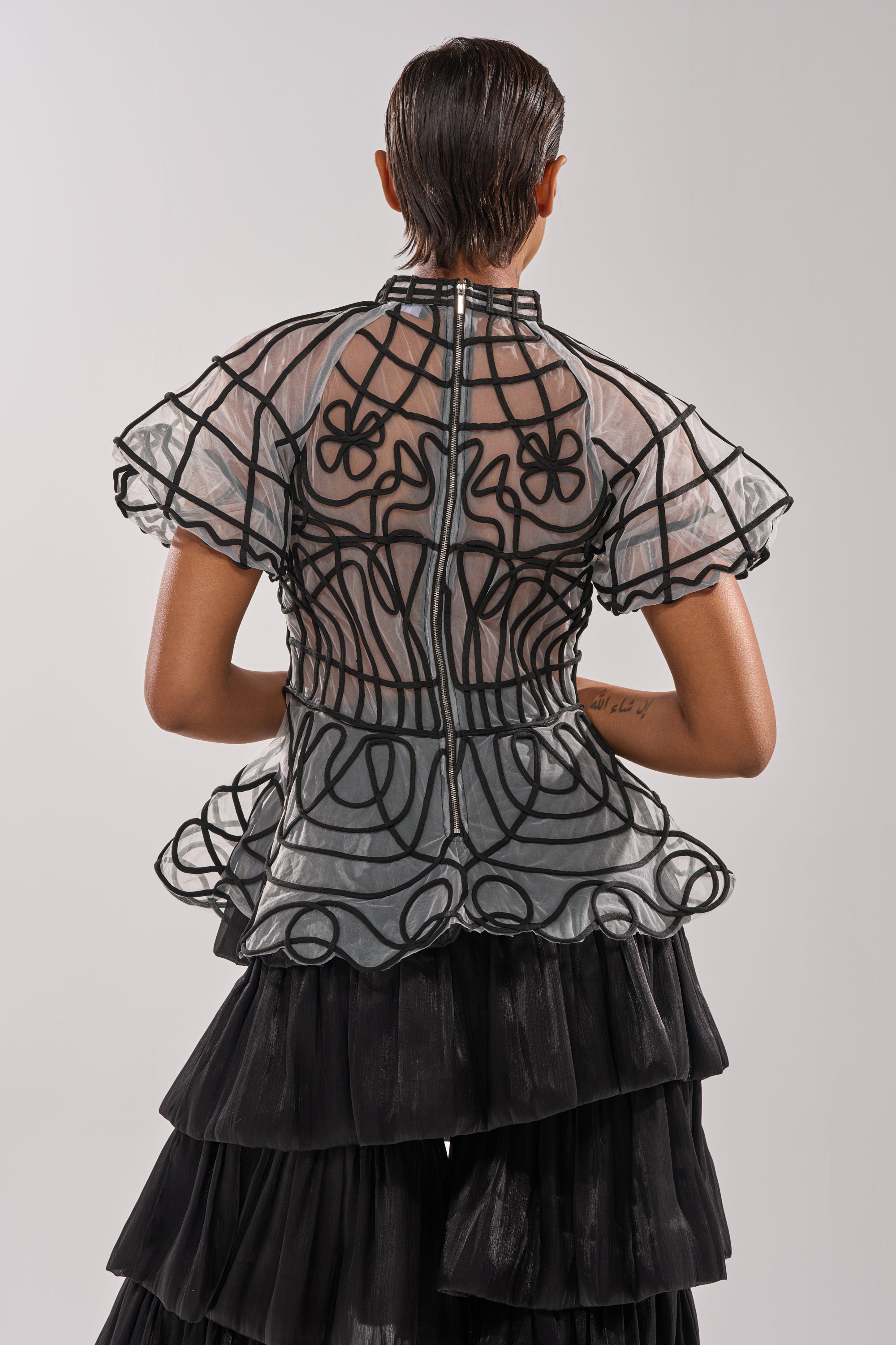 A person with short, slicked-back hair wears the DOLL FACE SHORT SLEEVE STRUCTURED BLOUSE featuring sheer mesh, black lines, and ruffles, paired with a tiered black skirt. The photo shows the blouse's back view against a plain background.