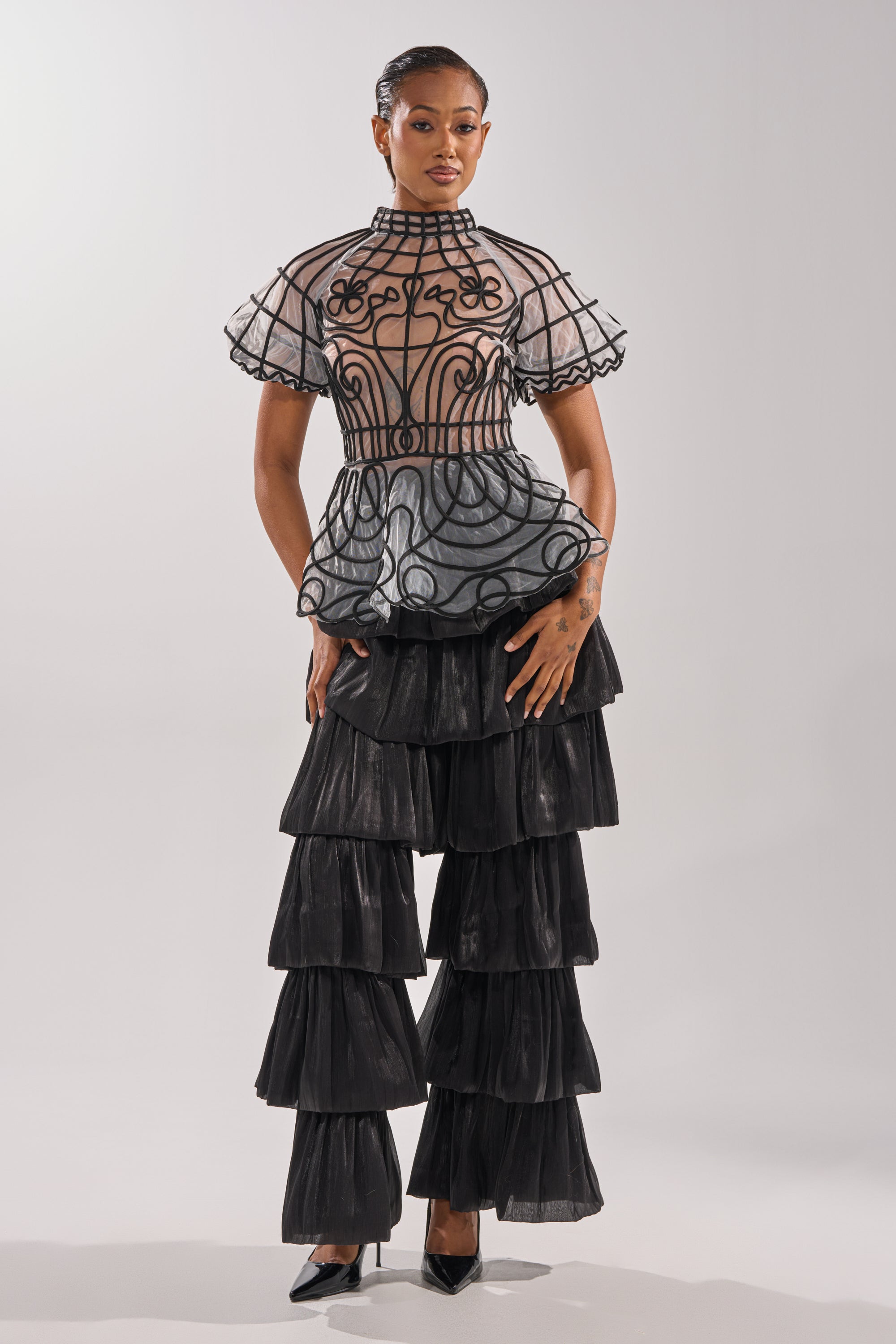 A woman models the DOLL FACE SHORT SLEEVE STRUCTURED BLOUSE, a sheer top with black swirls and ruffled sleeves, paired with tiered black ruffle pants and black heels against a plain background.