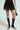 A person dons the SPIRITED AWAY SKIRT over sheer fabric, paired with knee-high, open-toe black boots and a small black handbag, set against a plain light grey background.
