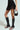Wearing the SPIRITED AWAY SKIRT, a person pairs it with black open-toe knee-high boots and holds a small black handbag in their right hand against a simple, light backdrop.