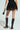 Wearing the stylish SPIRITED AWAY SKIRT with a high-rise silhouette, complemented by knee-high heeled boots and a small handbag, this modern outfit stands out against the plain background.