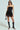 A woman stands confidently in a strapless black dress with a SPIRITED AWAY SKIRT featuring geometric cut-outs. She accessorizes with sunglasses, a small black handbag, and knee-high black heeled boots against a light background.