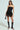 Wearing sunglasses, a person poses in the SPIRITED AWAY SKIRT, a black strapless dress with a structured lattice design and skater-style skirt. They hold a small black handbag and wear knee-high open-toe black boots against a plain backdrop.