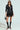 A person in a black leather jacket, a circular pattern SPIRITED AWAY SKIRT, knee-high black boots, and sunglasses holds a small black handbag against a plain background.