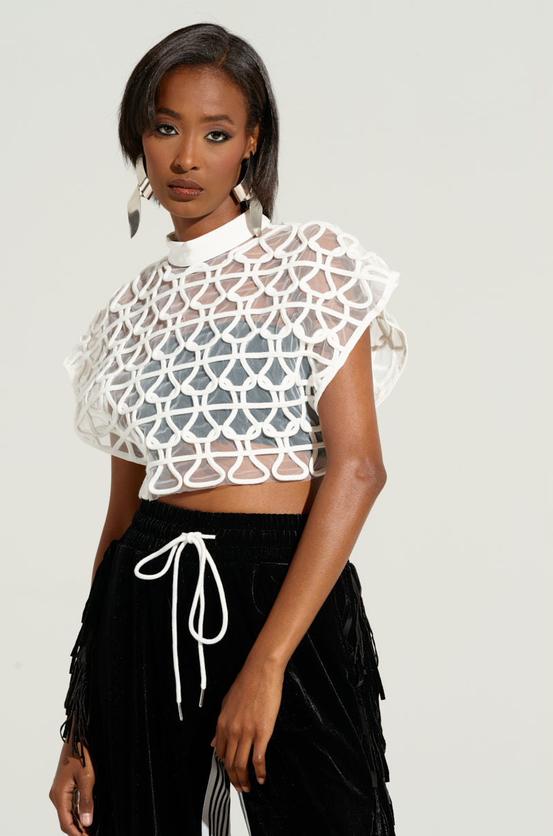 A woman is standing against a plain backdrop, clad in the PURE BLISS TOP—a sheer, geometric white crop top with abstract stripes overlaying a black top—paired with black velvet pants featuring a white drawstring. Her shoulder-length, straight hair complements her look, which is accented by large, silver, dangling earrings.
