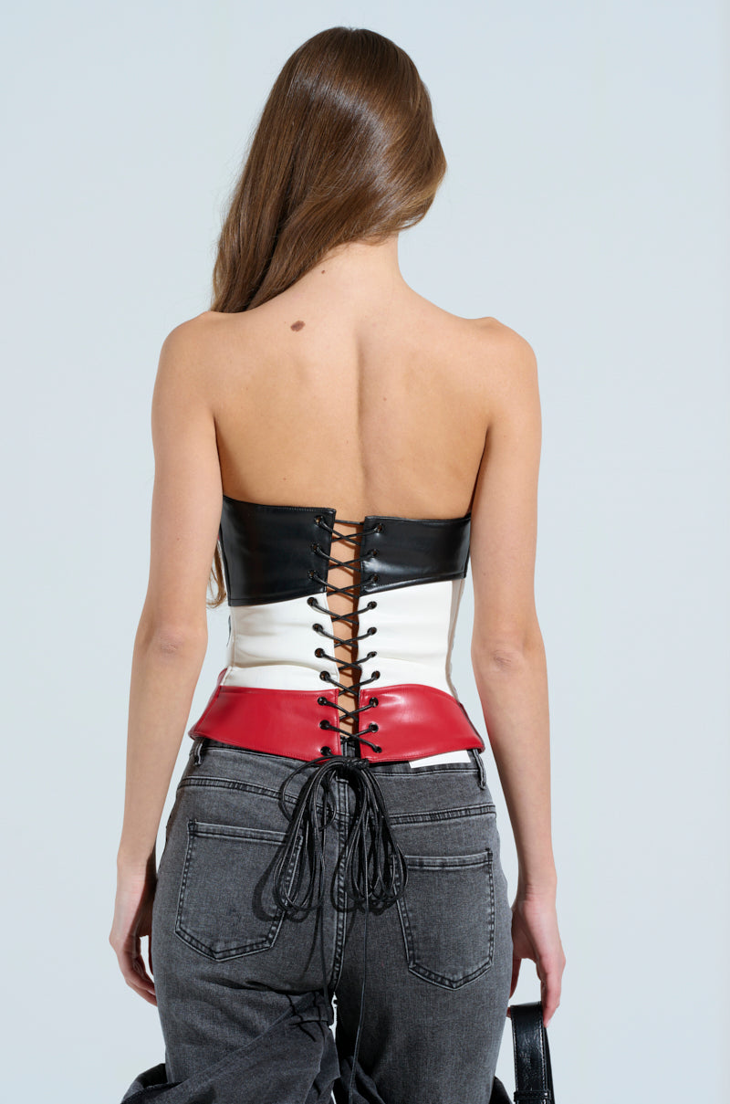 A woman with long brown hair, back to the camera, flaunts a waist-snatching silhouette in the MAKE ME FEEL CORSET. This strapless piece features black, white, and red hues with lace-up detailing. She pairs it with dark jeans and holds a handbag against a plain light gray background.