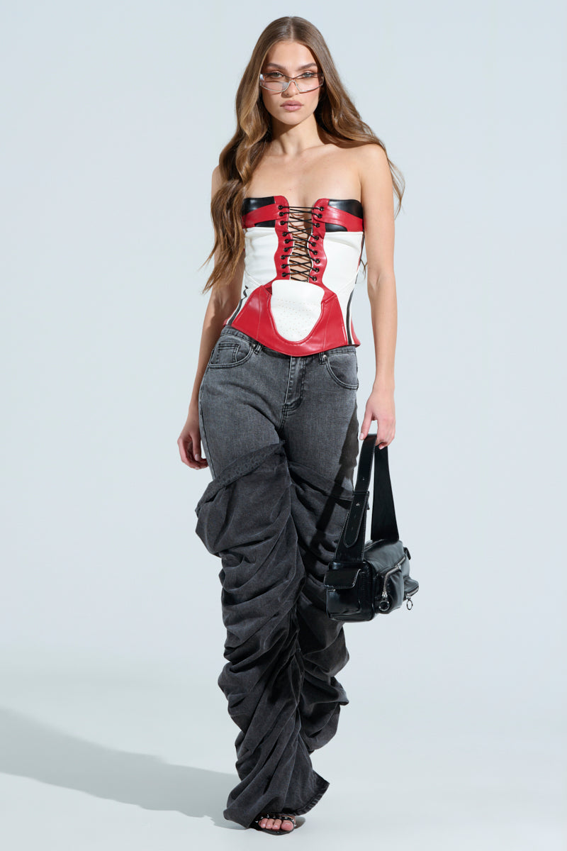 A woman with long hair wears sunglasses, a waist-snatching MAKE ME FEEL CORSET with lace-up detail, and dark ruched jeans. She carries a black handbag and stands against a plain background, effortlessly blending elegance and allure.