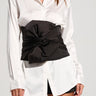 A person stands against a light background, wearing a white button-up shirt with the lower half open over black shorts. Their waist is cinched with the "Tied With A Bow Satin Belt," elegantly tied in a bow. The person has long straight hair and a necklace visible around their neck.