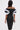 A woman with long dark hair wears the PRETTY WOMAN OMBRE MIDI DRESS, featuring a curve-hugging pencil silhouette and bold abstract ruffles on the off-the-shoulder sleeves. She holds a silver clutch, standing against a plain white background, facing away.