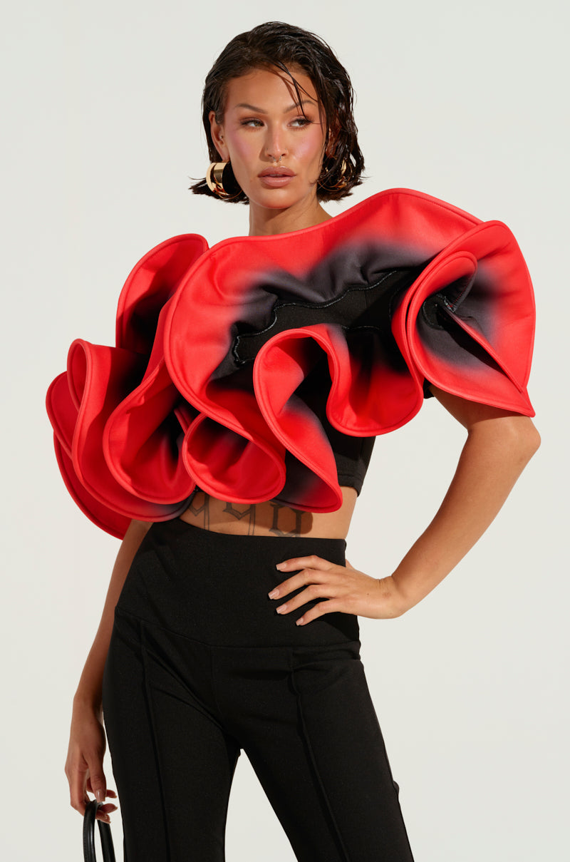 A person confidently models the PRETTY GIRL ROSE TOP, showcasing its avant-garde style with a striking red-and-black ombre design and dramatic ruffle detailing. They stand with one hand on their hip against a plain, light-colored background.