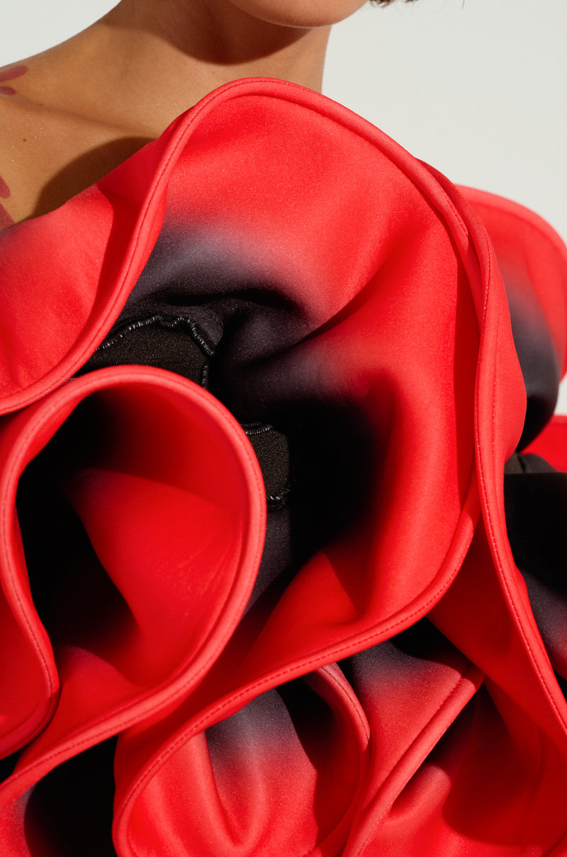 A close-up showcases the PRETTY GIRL ROSE TOP, a vibrant, abstract garment adorned with large, swirling red and black fabric ruffles that highlight its intricate detailing. The satin-like texture of the top creates dramatic folds and shadows, beautifully reminiscent of an ombre design against a plain, neutral background.