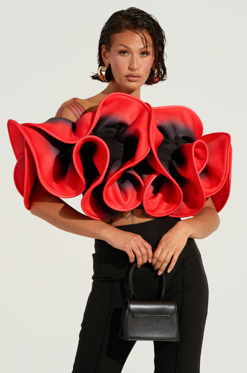 A woman showcases a striking fashion statement featuring the PRETTY GIRL ROSE TOP, with an eye-catching ombre design and layered red and black petals that resemble large roses. She accessorizes with a small black handbag, black pants, short wet-look hair, and large hoop earrings against a simple backdrop.