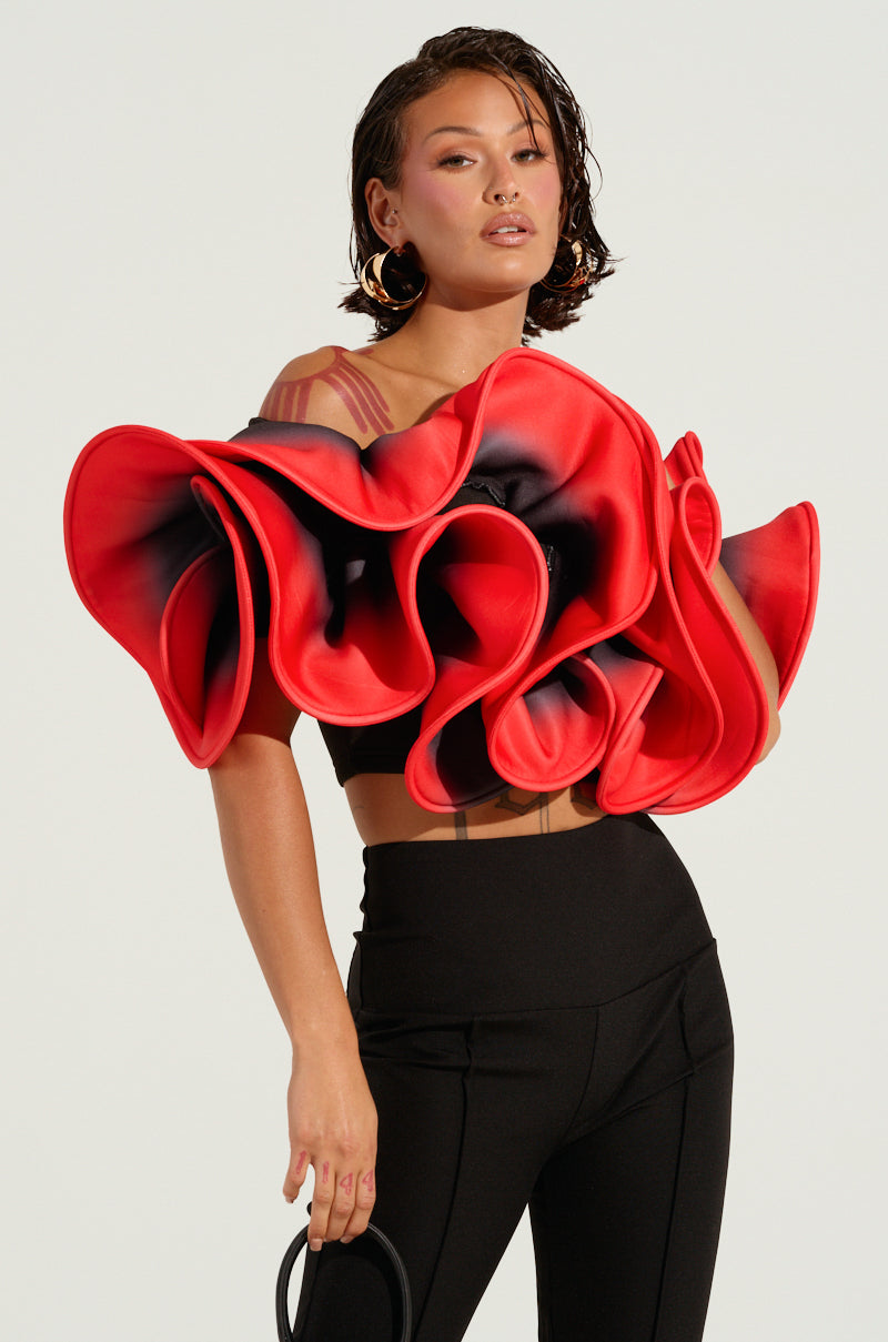 A person poses confidently, wearing the PRETTY GIRL ROSE TOP with its bold, sculptural rose design and dramatic ruffle detailing, paired with sleek black pants. The visually striking outfit resembles a flower in bloom against a simple, light-colored background.