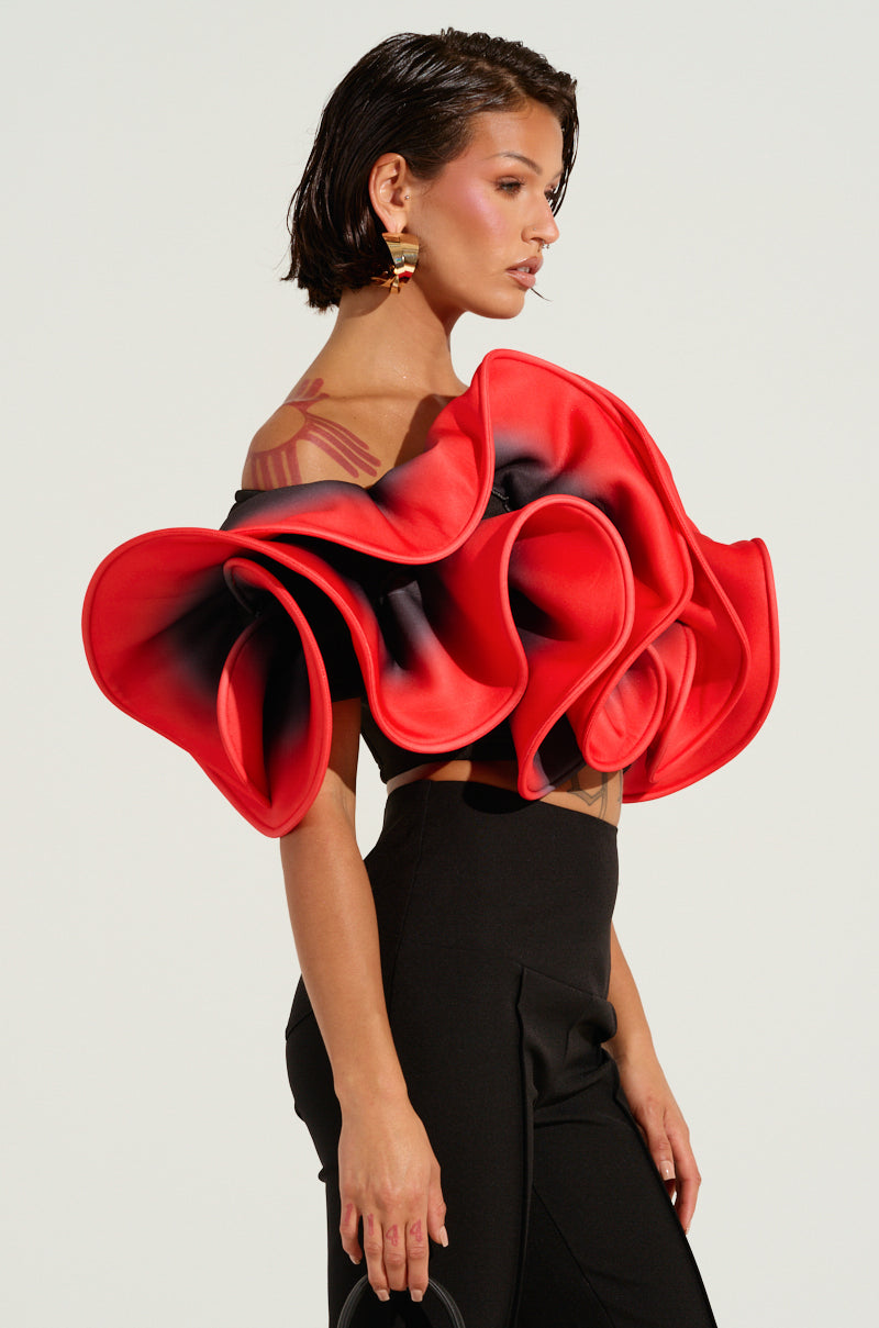 A person with short dark hair is wearing the PRETTY GIRL ROSE TOP paired with black pants. The ruffle detailing enhances their style as they glance to the side, highlighting gold earrings and a tattoo on their shoulder against a plain light background.