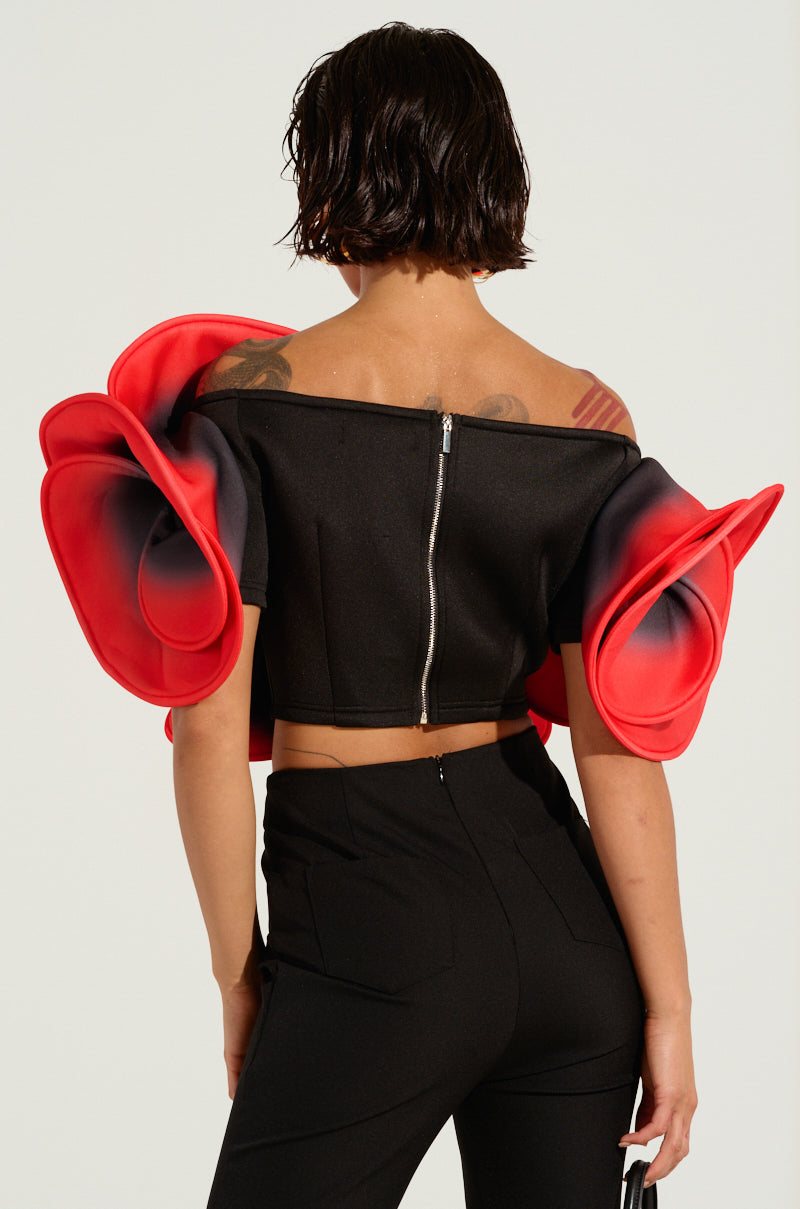 A person with short, dark hair models the PRETTY GIRL ROSE TOP, a black off-shoulder garment adorned with large red floral sleeves and matched with black pants. They face away to showcase the zippered back of the top, which is complemented by a subtle ombre design against a simple background.
