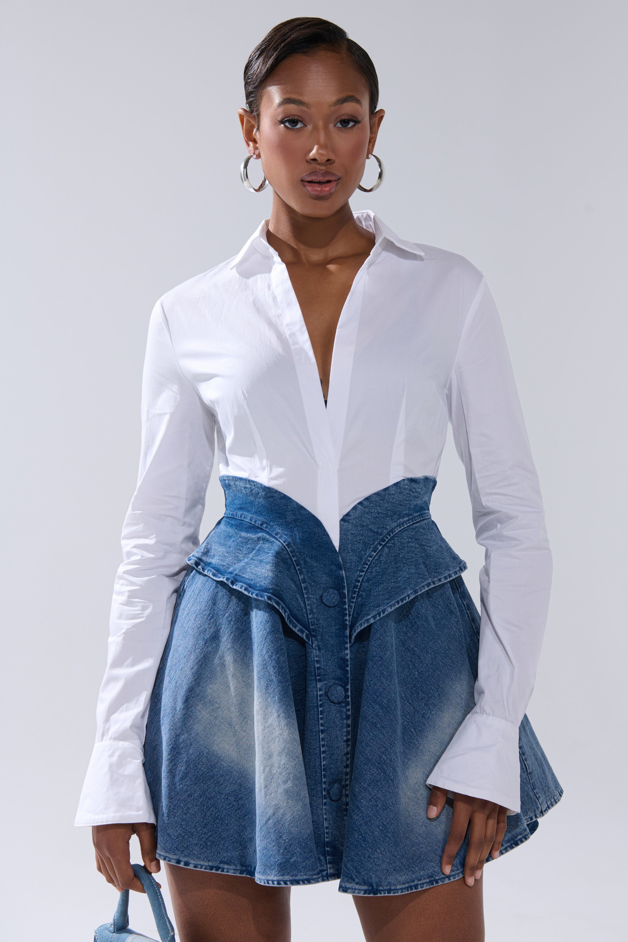 A woman poses confidently against a neutral background wearing the DONT NEED NOBODY DENIM MINI DRESS, featuring high-waisted design, large buttons, and paired with a long-sleeved blouse.