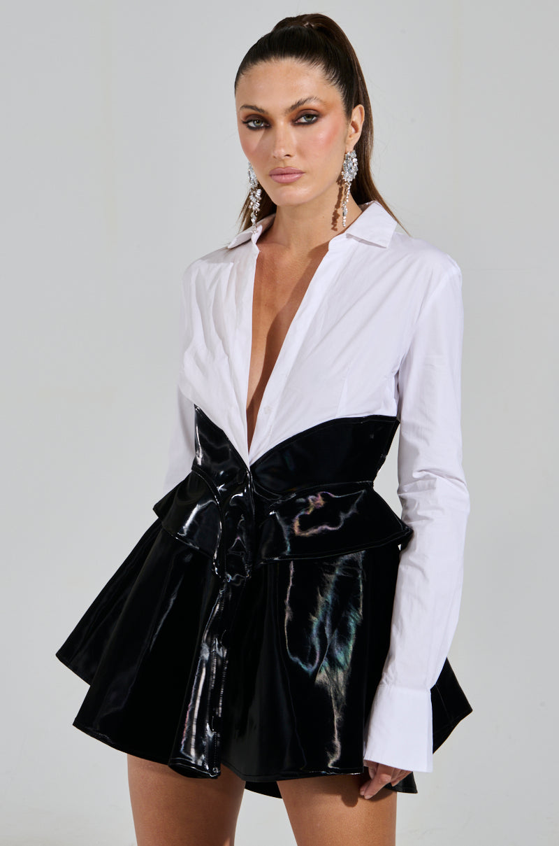 A person in a MODERN ELEGANCE POPLIN MINI DRESS with a black, glossy finish and an open-neck white shirt underneath. Their long hair is tied back, and they're accessorizing with large earrings. The backdrop is plain gray.