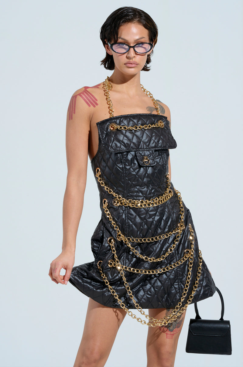Dressed in the stylish GOOD VIBES Quilted Nylon Mini Dress highlighted by metallic chain links, a confident individual poses with glasses and a small black handbag. Their tattoos are visible on their arms and shoulder as they stand against a light background.