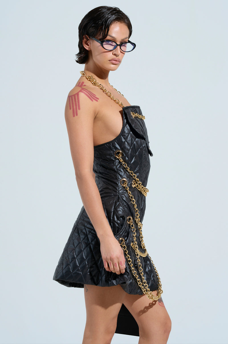 A person with short dark hair and glasses wears a stylish black GOOD VIBES QUILTED NYLON MINI DRESS, enhanced with metallic chain links. They feature a red design on their shoulder and project confidence against the plain background.