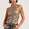 A person stands confidently against a pale backdrop, showcasing the HIGH END RODEO TURQUOISE STONE TOP, which features Western-inspired designs and intricate patterns. They sport short dark hair, large hoop earrings, visible arm tattoos, and are paired with vintage-inspired blue jeans.