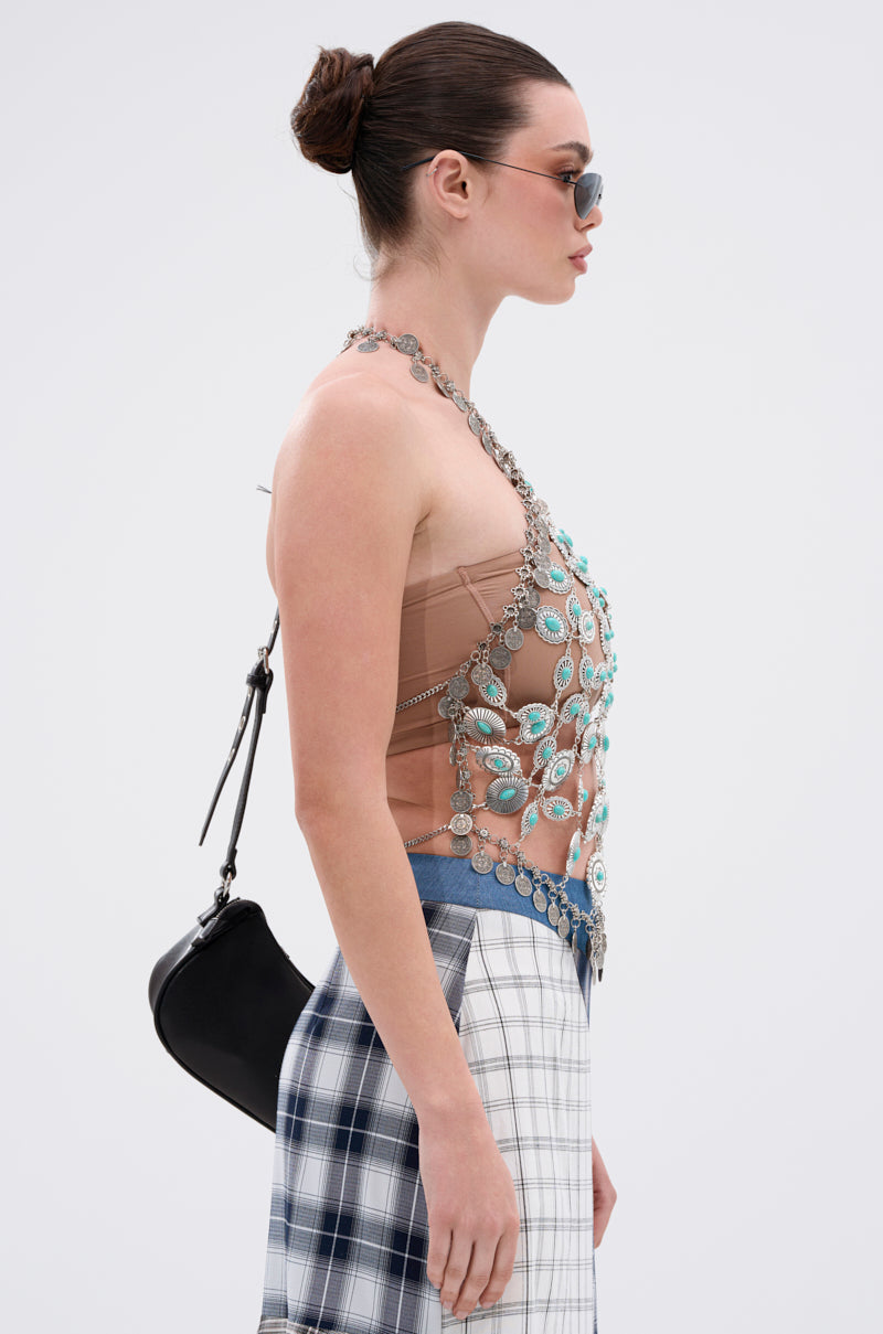 A woman in profile wears sunglasses, the HIGH END RODEO TURQUOISE STONE TOP with silver accents, a silver chain necklace, plaid skirt, carries a black shoulder bag, and has her hair styled in a bun.