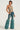 The person confidently wears the HIGH END RODEO TURQUOISE STONE TOP, a sheer, vintage-inspired garment with circular embellishments, paired with blue jeans featuring large fuzzy boot covers extending from the knees to the ankles. The background is a plain off-white.