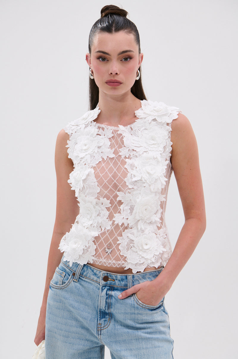 A woman wears the UNTAMED FLORAL LACE TOP, a sleeveless sheer mesh piece with white 3D floral appliqués, paired with light blue jeans. Her hair is in a high ponytail, and she poses with one hand in her pocket against a plain background. 0