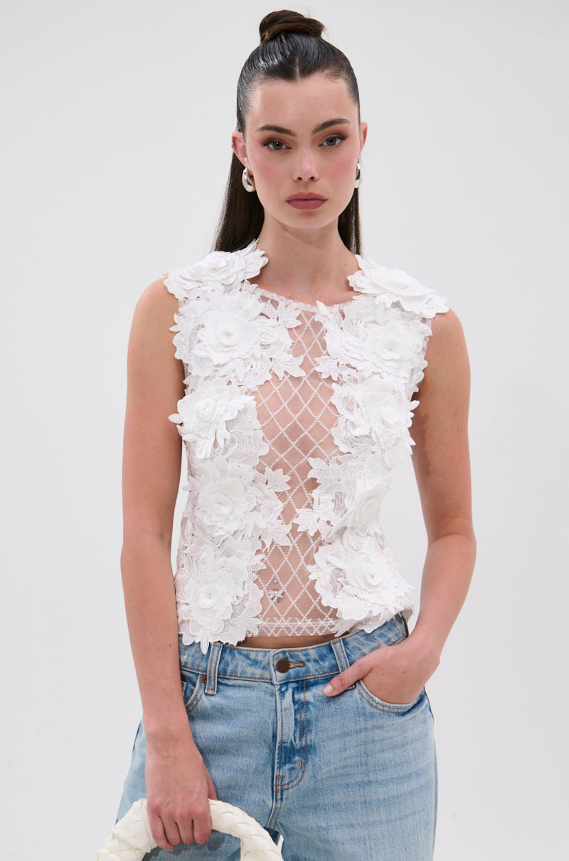 A woman with long dark hair in a high ponytail wears the UNTAMED FLORAL LACE TOP—sleeveless, white, with 3D floral appliqués and a sheer mesh panel—paired with light blue jeans against a plain white background. 1