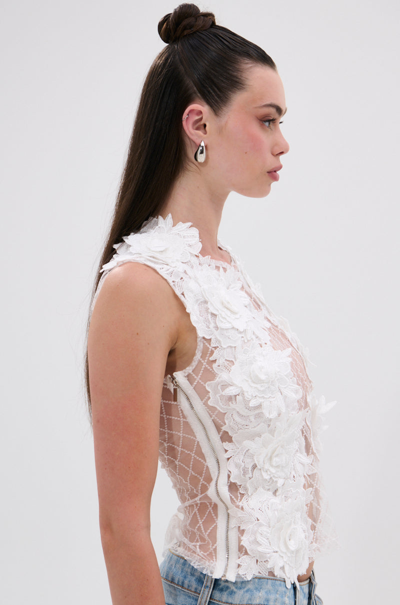 A woman in profile wears the UNTAMED FLORAL LACE TOP, a sheer mesh white blouse with 3D floral details and geometric patterns, paired with light blue jeans. Her long dark hair is styled half-up, and she wears geometric earrings. 2