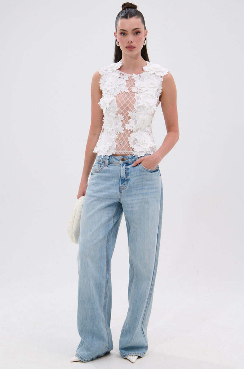 A woman with long dark hair pulled back models the UNTAMED FLORAL LACE TOP—a sleeveless white lace top with mesh accents—paired with light blue high-waisted, wide-leg jeans and a white textured bag against a plain backdrop. 4