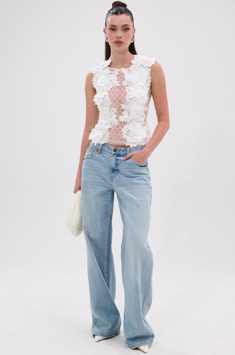 A woman poses against a white background in the UNTAMED FLORAL LACE TOP with 3D appliqués, light blue wide-leg jeans, a white textured clutch, pointed-toe shoes, and her hair tied back. 5