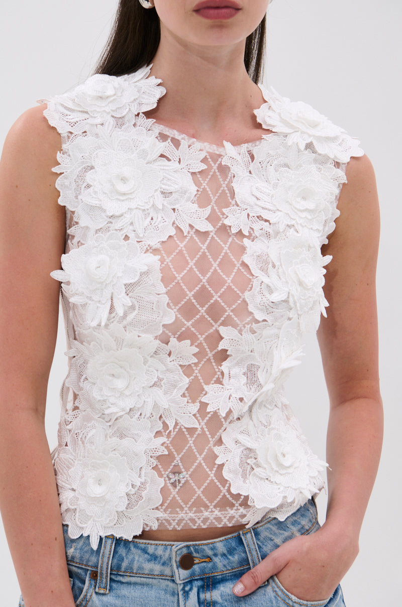 A woman wears the UNTAMED FLORAL LACE TOP—a sleeveless, sheer mesh top with white 3D floral appliqués and a crisscross pattern—paired with light blue jeans against a plain, light background. 6