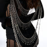 A person in a black, sheer outfit is viewed from behind, with long, wavy hair cascading down. Multiple layers of silver chains and glittering rhinestone embellishments from the AKIRA Label’s FOR THE MET GLOVE BOLERO drape across their back, creating an ornate and elegant design. The interplay of sheer and opaque sections adds to its sophisticated allure.