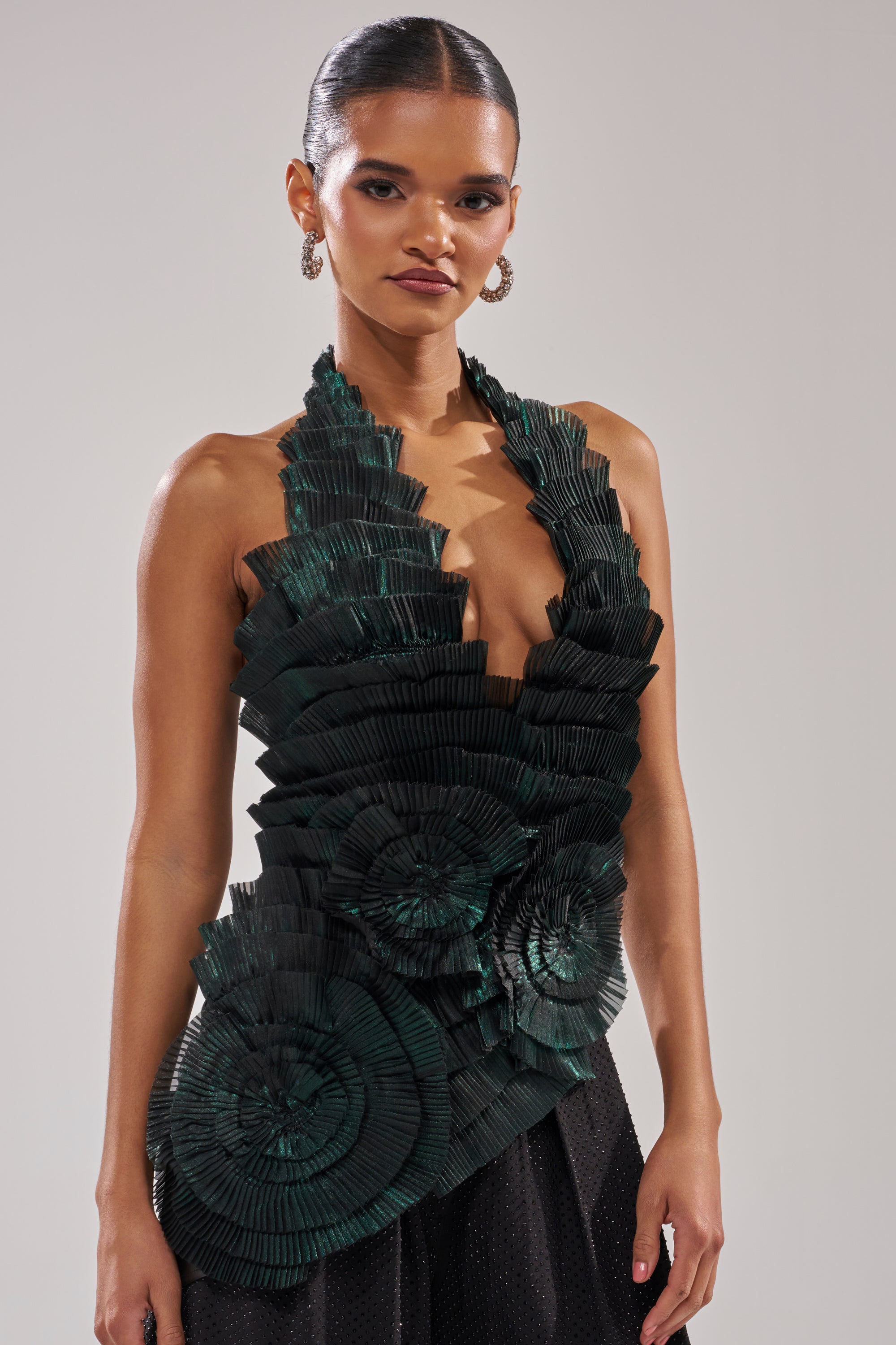 A woman with slicked-back hair models the DREAMERS HALTER BLOUSE in emerald green, showcasing layered fan-like pleats and circular floral motifs, paired with black pants against a neutral background.