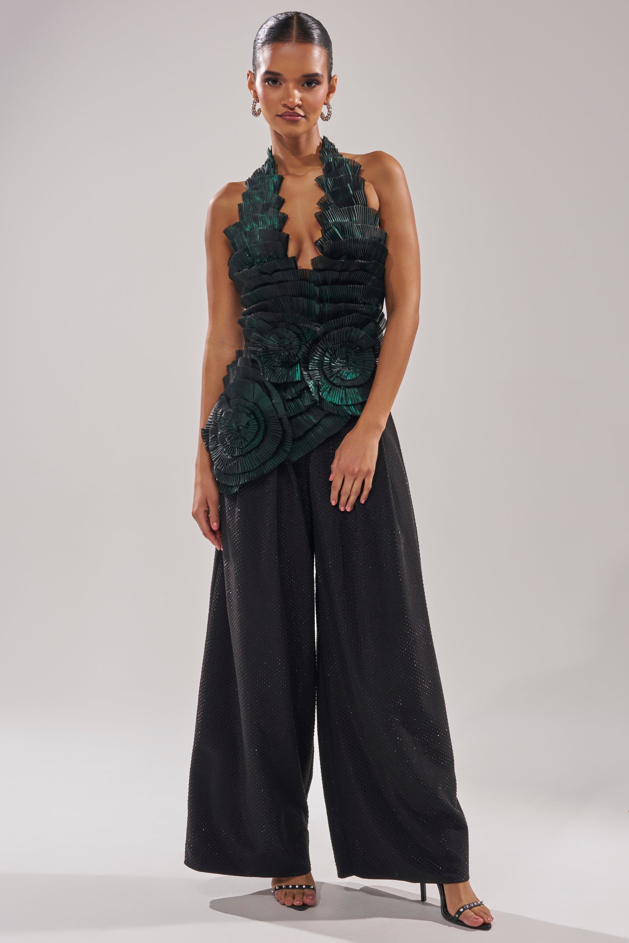 Wearing the DREAMERS HALTER BLOUSE in emerald green with bold leaf-like details, a woman poses against a plain background. She pairs it with black wide-leg pants, hoop earrings, slicked-back hair, and black heeled sandals.